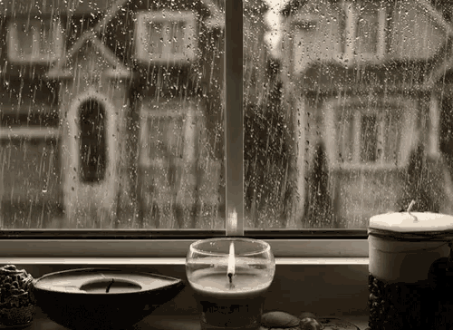 a candle is lit in front of a window with rain drops on it and a house in the background .