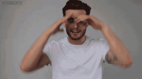 a man in a white t-shirt is covering his eyes with his hands and smiling .