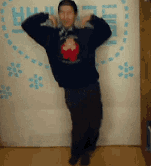 a man in a black sweater with a red heart on it is standing in front of a wall with flowers on it .