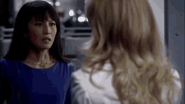 two women are standing next to each other and talking to each other . one of the women is wearing a blue dress .