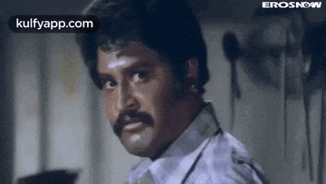 a man with a mustache and a white shirt is looking at the camera in a room .
