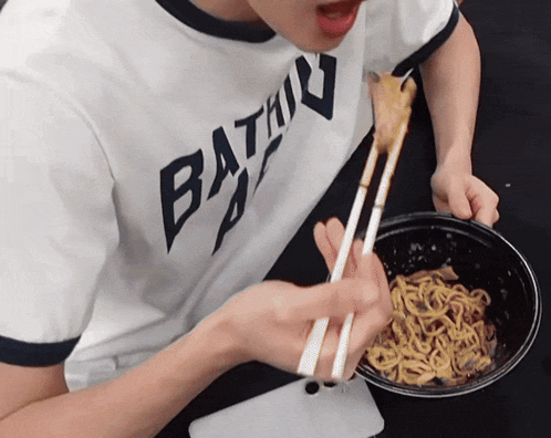 a person in a bathing ape shirt is eating noodles with chopsticks