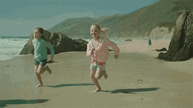 two young girls are running on a beach and one has a pink jacket on