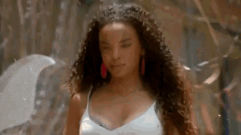 a woman with curly hair wearing a white tank top and red earrings is standing in front of a bubble .