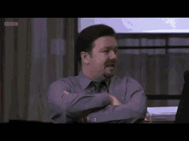 a man in a purple shirt is sitting at a desk with his arms crossed in front of a bbc logo