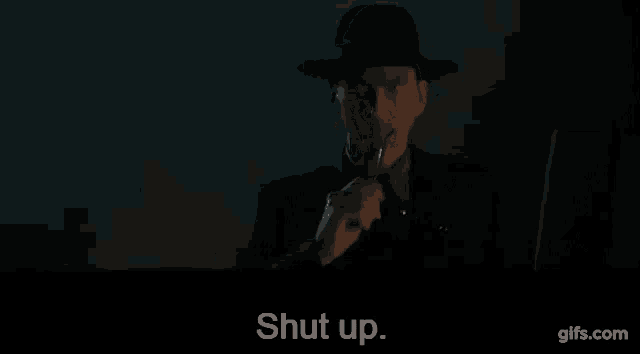 a man in a suit and hat is holding a cigarette in his mouth and says `` shut up '' .