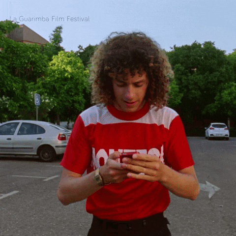 a man with curly hair wearing a red shirt that says la guarimba film festival is looking at his phone