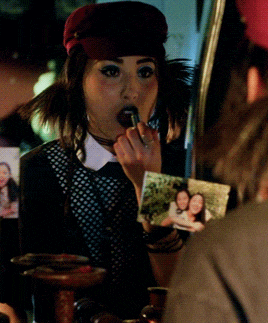 a woman is applying lipstick in front of a mirror with a picture of two women behind her