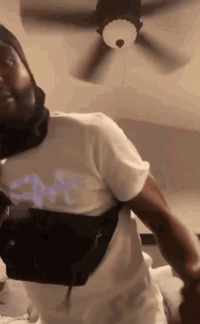 a man wearing a white t-shirt with a black bag around his waist is standing under a ceiling fan