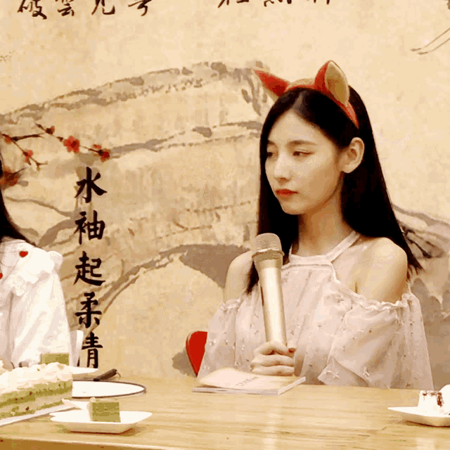 a woman sitting at a table holding a microphone in front of a wall with chinese writing on it