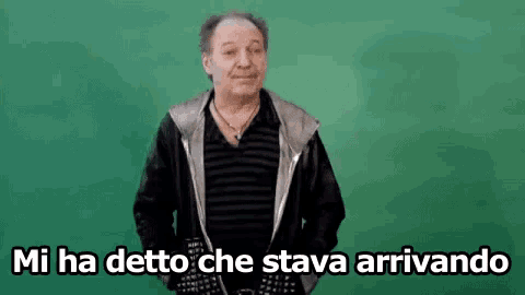 a man in a black jacket is standing in front of a green background and says `` mi ha detto che stava arrivando '' .