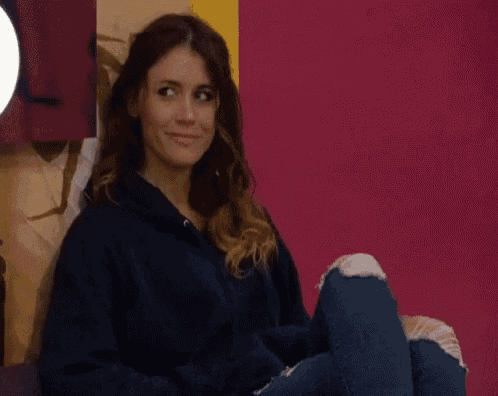 a woman is sitting in front of a pink wall with her legs crossed and making a funny face .