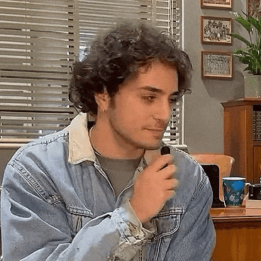 a man in a denim jacket is sitting at a desk in front of a window in a room .