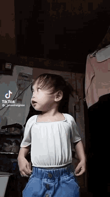 a little girl in a white shirt and blue jeans is standing in a dark room .