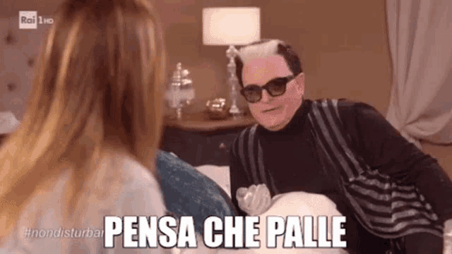 a man wearing sunglasses is sitting on a bed talking to a woman with the words pensa che palle below him