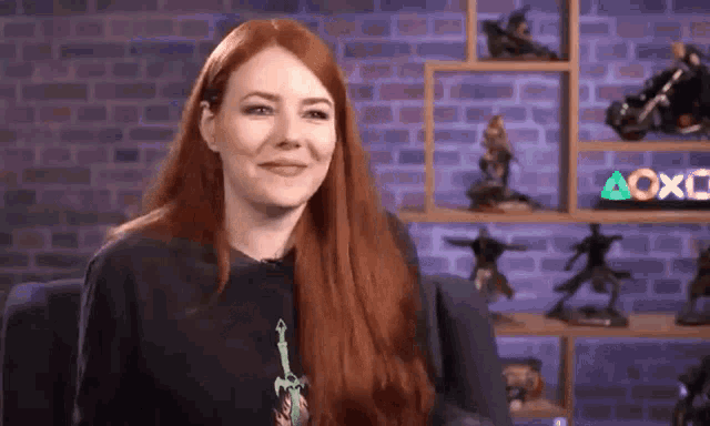 a woman with long red hair is sitting in front of a brick wall and a shelf filled with figurines .