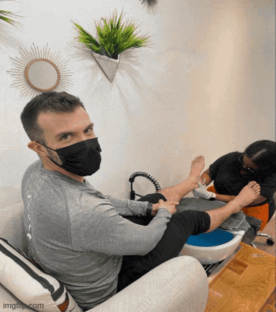 a man wearing a face mask sits on a couch while a woman does a manicure