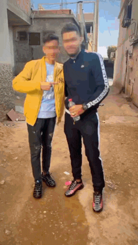 two men are standing next to each other on a dirt road . one is holding a bottle of water .