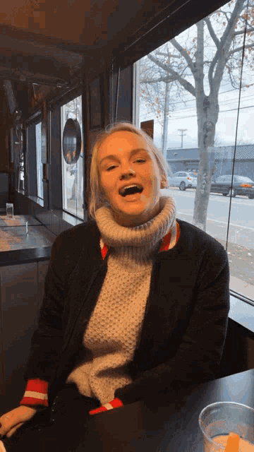 a woman wearing a turtleneck sweater and a black jacket sits at a table with her mouth open