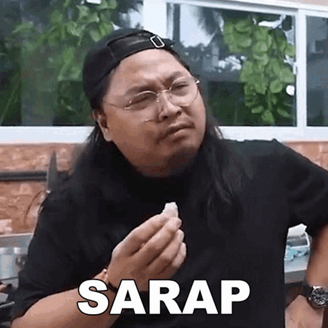 a man with long hair and glasses is holding a piece of food in his hand and the word sarap is above him .