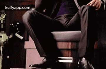 a man in a suit is sitting in a chair with his legs crossed and talking on a cell phone .