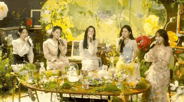 a group of women are singing into microphones while sitting around a table surrounded by flowers .