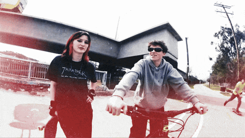 a girl wearing a thrasher shirt is standing next to a man riding a bike