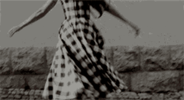 a woman in a plaid dress is dancing in front of a stone wall in a black and white photo .