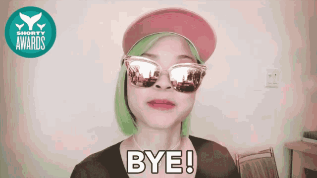 a woman with green hair wearing sunglasses and a pink hat says bye