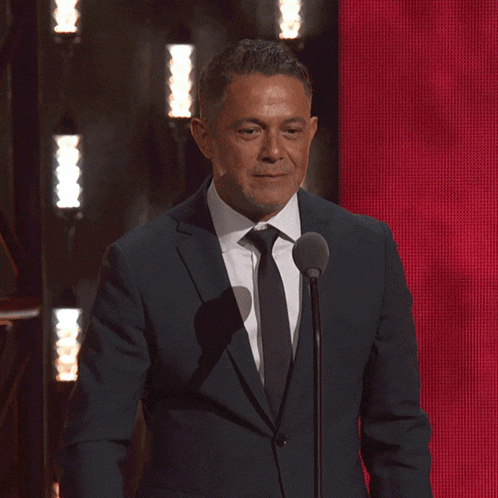a man in a suit and tie is pointing at the camera while speaking into a microphone