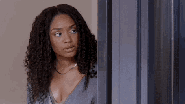 a woman with curly hair is standing next to a door and looking at the camera .