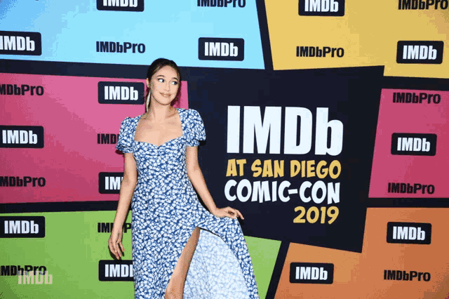 a woman in a blue dress stands in front of a wall that says imdb at san diego comic-con 2019