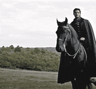 a man in a cape rides a black horse in a field