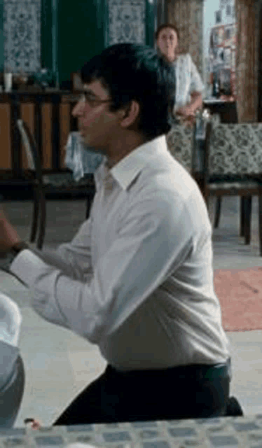 a man in a white shirt is kneeling down in a room with a woman in the background .