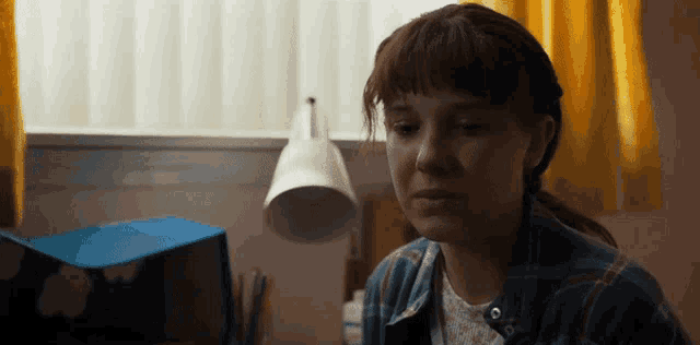 a girl in a plaid shirt sits in a room with a lamp and a blue stool
