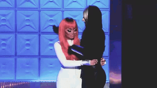 two women are hugging each other in front of a blue wall . one of the women is wearing a red wig .