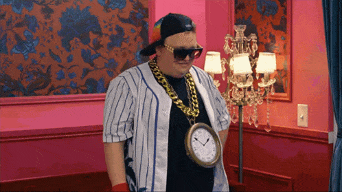 a man wearing sunglasses and a clock around his neck is standing in front of a chandelier