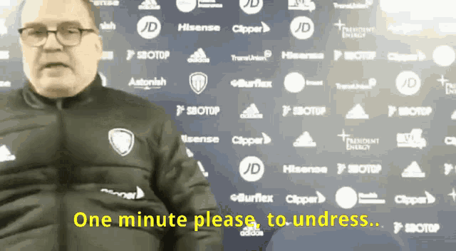 a man is standing in front of a wall of logos and says " one minute please "