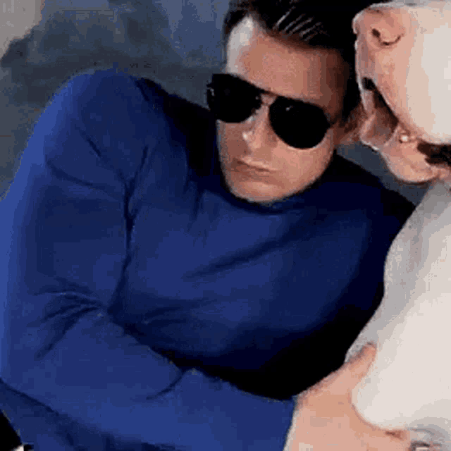 a man wearing sunglasses and a blue sweater is kissing another man on the cheek .
