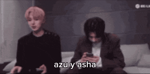 two men are sitting on a couch looking at their phones and one of them is saying `` azu y asha '' .
