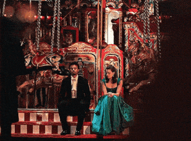 a man in a suit sits next to a woman in a blue dress in front of a merry go round