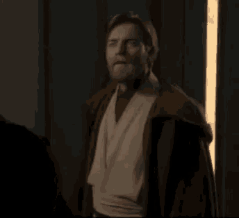 a man in a brown robe is standing in a dark room with a light behind him .