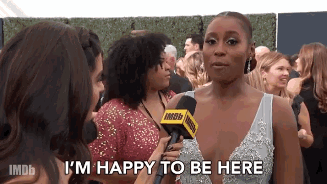 a woman in a dress is talking into a microphone and saying i 'm happy to be here .