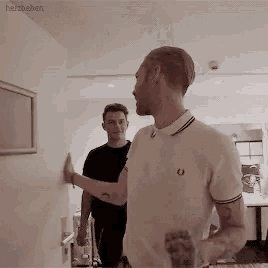 two men are standing next to each other in a room . one of the men is leaning against a wall .