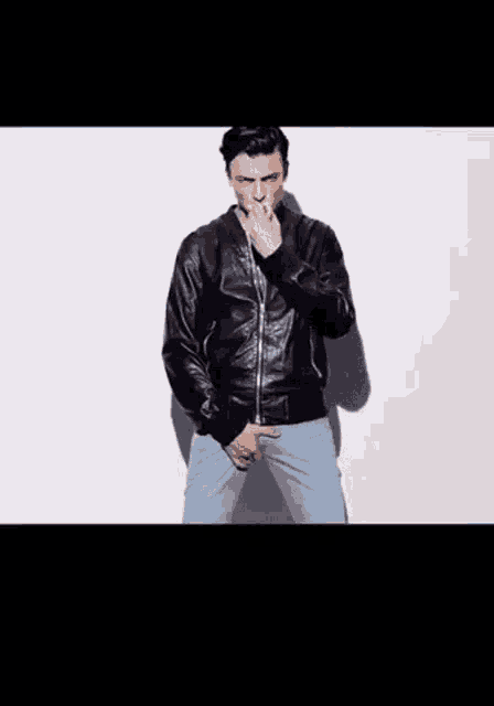 a man in a black leather jacket and blue jeans is standing in front of a white wall .