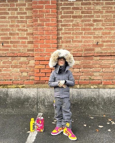 a child wearing a fur hood stands in front of a brick wall next to a bag of bubbles