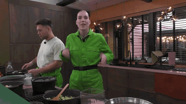 a man and a woman are cooking in a kitchen and the woman is wearing a green jumpsuit
