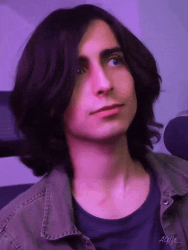 a young man with long dark hair and blue eyes is wearing a purple shirt and a green jacket .