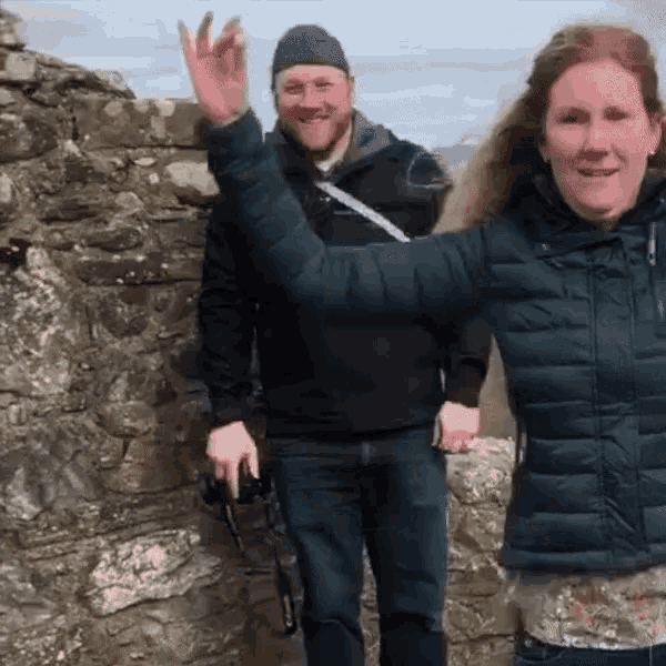 a man and a woman are standing next to a stone wall and the woman is waving
