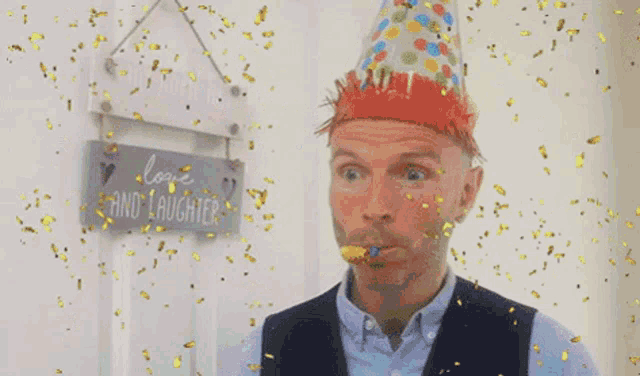 a man wearing a party hat is blowing out a party horn while confetti falls around him .